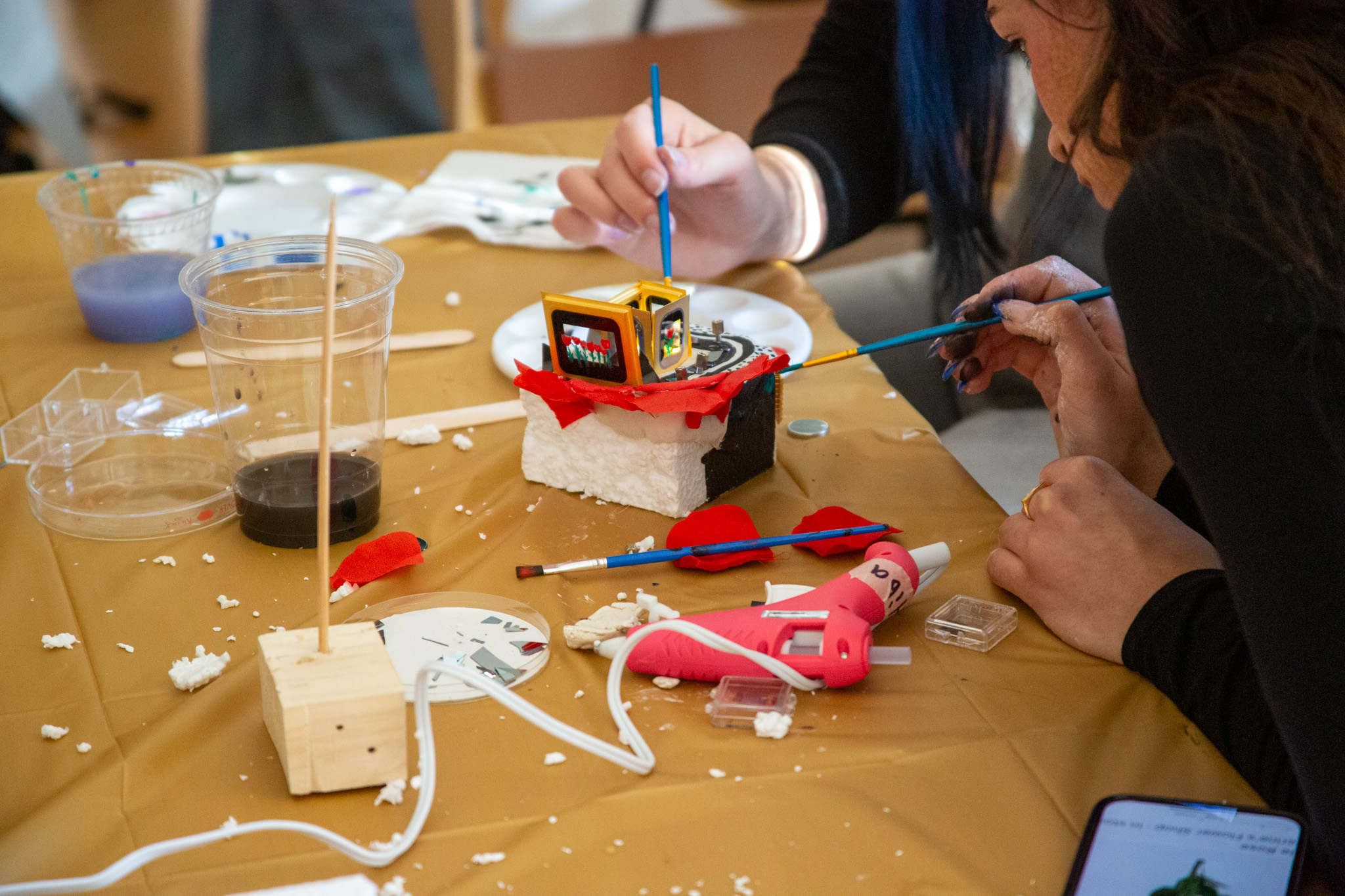 Detailed view of a mixed‑media sculpture created during the “Cosmic Missteps” workshop, showing a small cube mounted on a foam base and accented with paint and structural elements, while hands nearby apply finishing touches using brushes and tools.