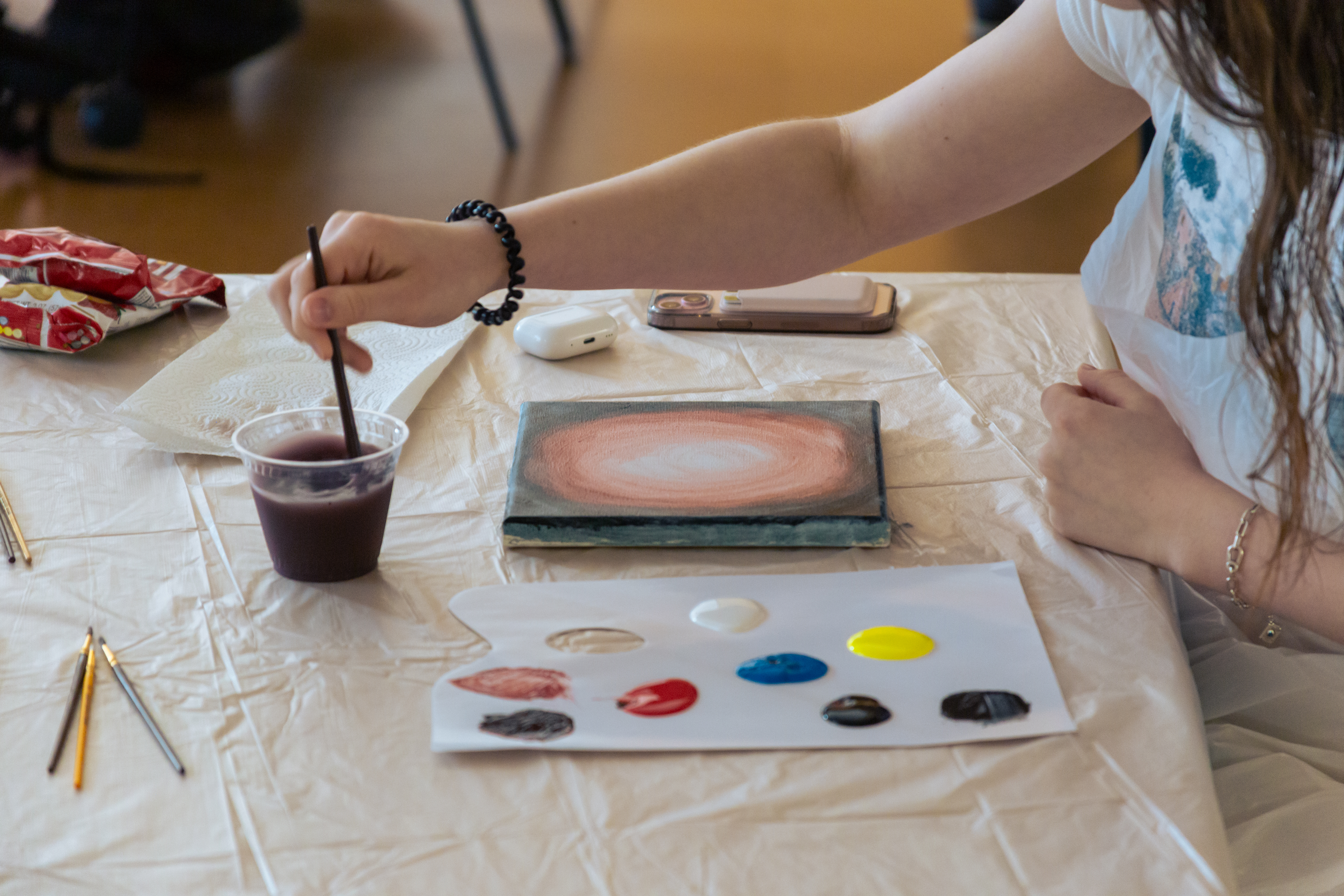 During a Cosmic Missteps event, a participant paints at a table with acrylic colors laid out on a palette, a small canvas in progress, paint brushes, and personal items arranged on a protective table covering.